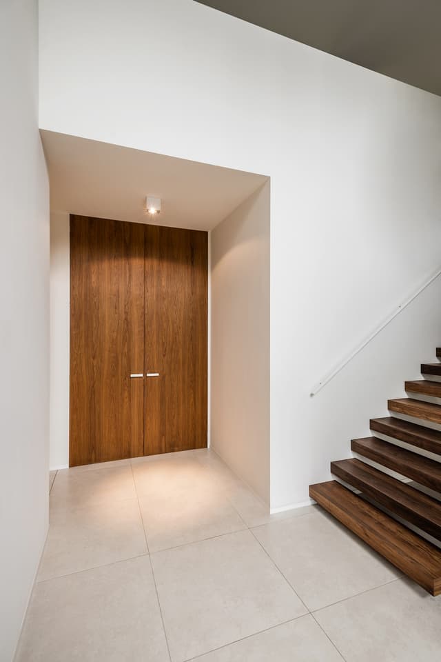 A tiled hallway with wooden double flush doors and a brown floating stairs leading out of the frame to the right, white walls and a set of closed double flush brown wooden doors with silver handles.